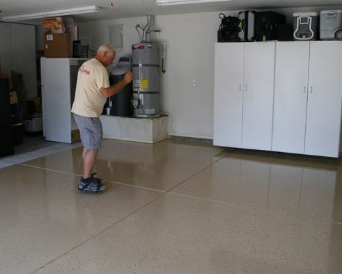 The image shows men painting a floor using a roller brush. He is painting a garage floor.